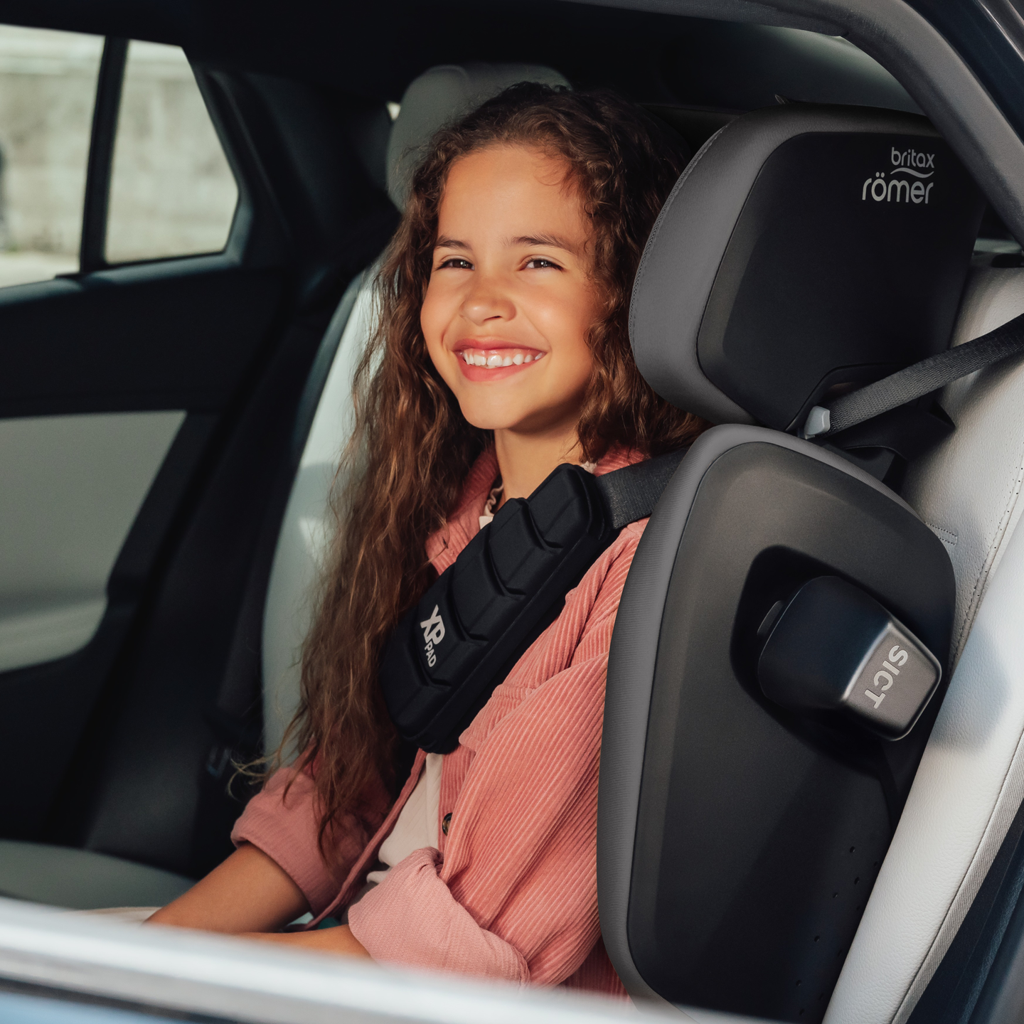 Uśmiechnięte dziecko na tylnym siedzeniu samochodu, przypięte w foteliku samochodowym Britax R&ouml;mer KIDFIX PRO z wysokim oparciem.